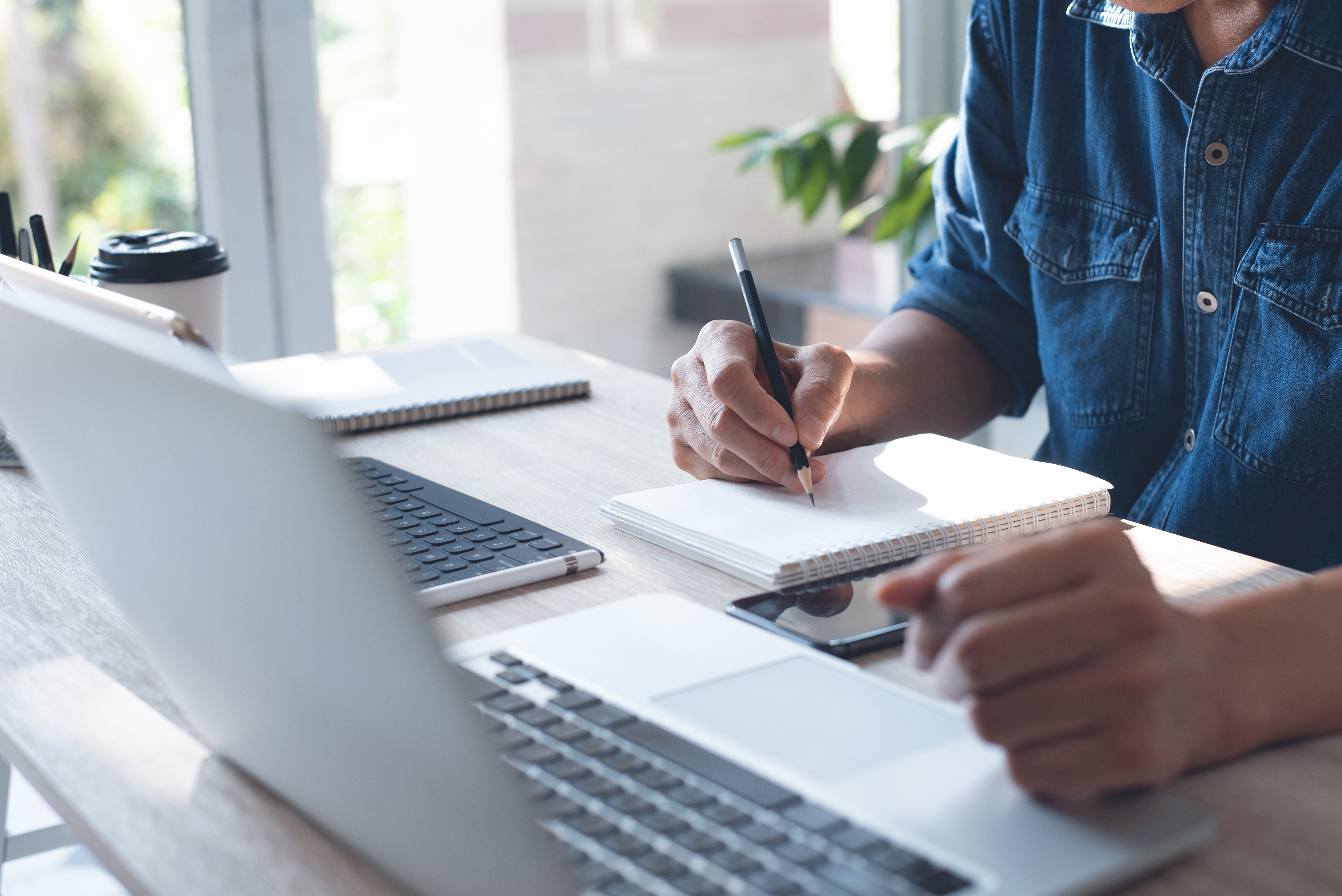 Close-up of a man writing in a notebook while working on a laptop.