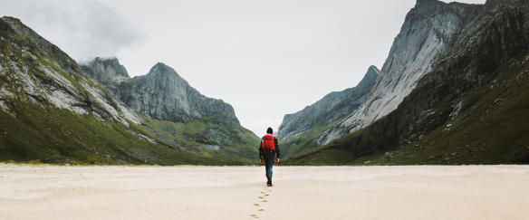 Man with a backpacks walks alone in the sand towards a lush mountain valley