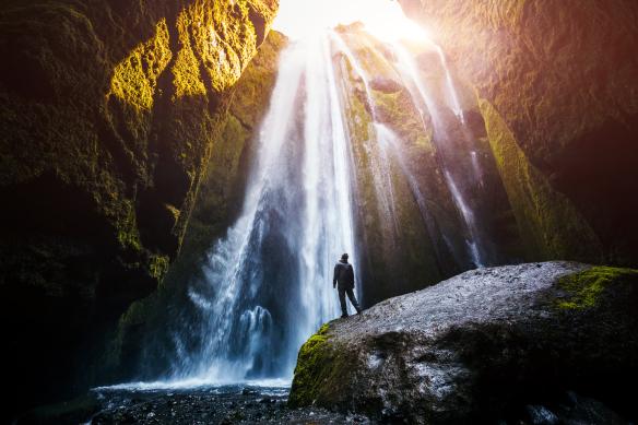 A person in silhouette looks up at a waterfall cascading down with the sunlight shining down
