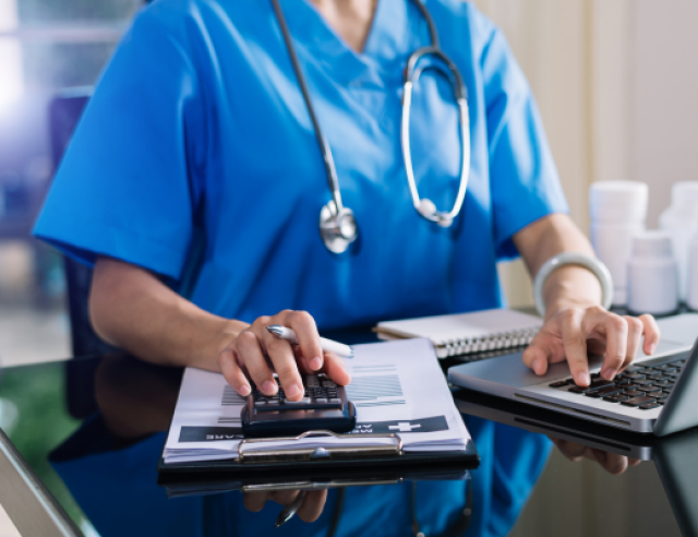 A person wearing blue medical scrubs with a stethoscope around their neck, sitting at a desk. They are using a calculator with one hand and typing on a laptop with the other. On the desk, there is a clipboard with papers, a pen, and some medication bottles in the background. The setting suggests a healthcare professional performing administrative or financial tasks.