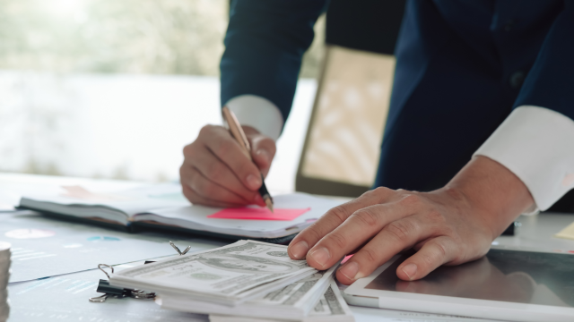 A close-up of a person wearing a suit, writing on a notepad with a pen. Their other hand rests on a stack of hundred-dollar bills placed on a table alongside various papers and a tablet.