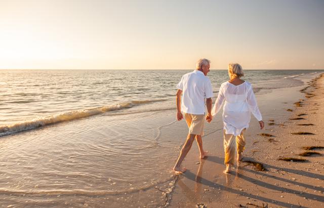 A happy senior couple holds hand while walking down a beach at sunrise