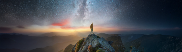 A man stands atop a mountain with his arm raised. A starry sky fills the background at sunrise. 