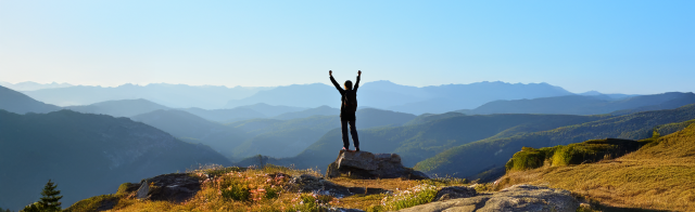 A woman stands with her arms raised at the top of a mountain looking over a dense forest valley.
