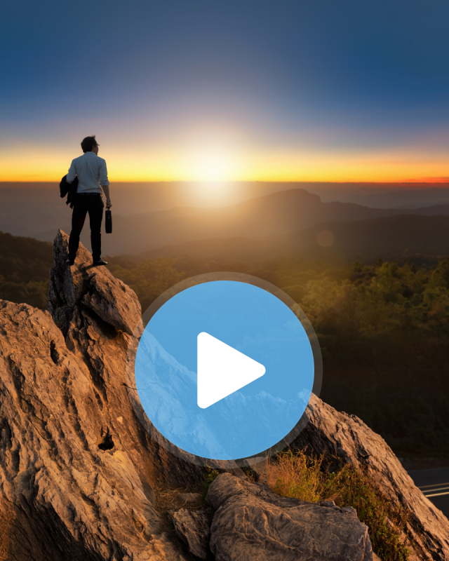A businessman stands atop a mountain peak holding a briefcase and looking out at the sunset